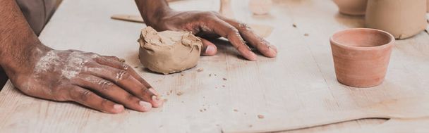 vista parcial del joven afroamericano con las manos cerca de un pedazo de arcilla sobre la mesa con equipo en cerámica, pancarta - Foto, Imagen