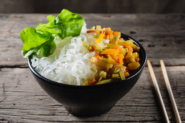 Un plato de fideos de arroz se encuentra sobre una vieja mesa de madera. Los fideos de arroz son más comunes en las cocinas del este, sudeste de Asia y sur de Asia. El guarnición consiste en cebollas fritas, zanahorias, pimentón. Copiar espacio. Orientación horizontal - Foto, Imagen