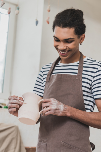 smiling young african american man sculpting clay pot with stick in pottery - Foto, Imagen