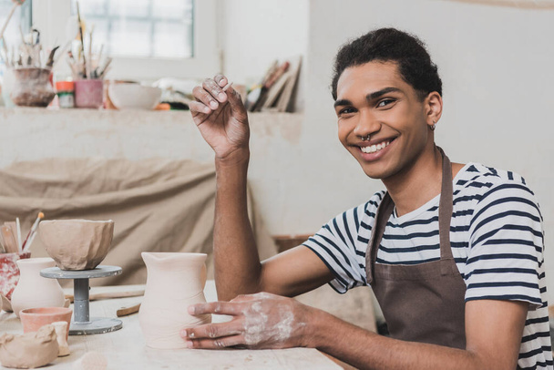 smiling young african american man looking at camera near clay pot and stick in hand in pottery - Photo, Image