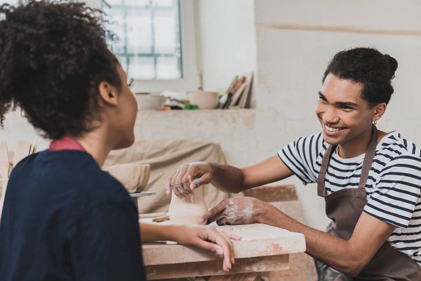 улыбающийся молодой африканский американец, создающий глиняный горшок и палку и смотрящий на женщину поблизости в керамике - Фото, изображение