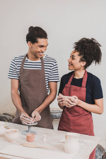 smiling young african american couple in aprons making clay pots and looking at each other in pottery - Photo, Image