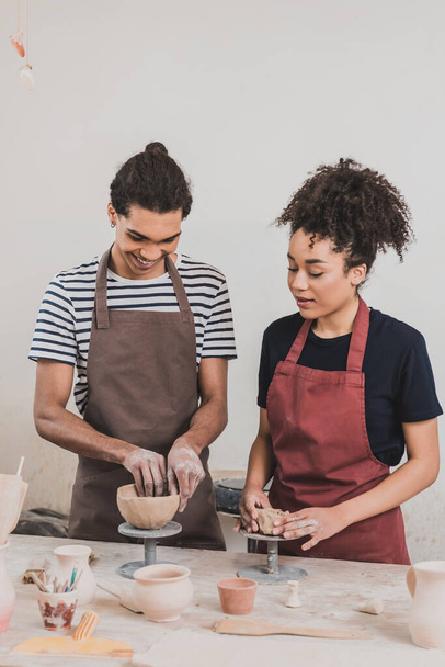 alegre joven afroamericano pareja en delantales haciendo ollas de barro en la mesa con equipo en cerámica - Foto, Imagen