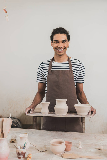 smiling young african american man standing with clay pots on tray in pottery - Photo, Image