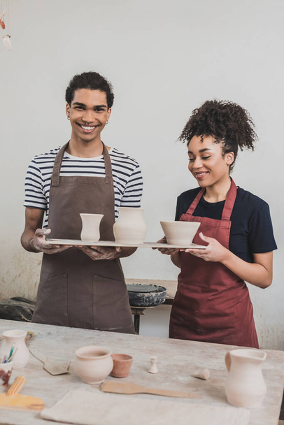 joven pareja afroamericana positiva sosteniendo ollas de arcilla en bandeja en cerámica - Foto, imagen
