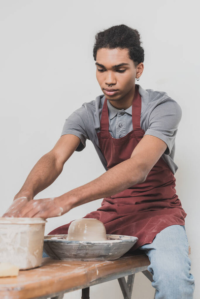 serieuze jonge Afrikaanse Amerikaanse man wassen handen in plastic doos in de buurt natte klei op wiel in aardewerk - Foto, afbeelding