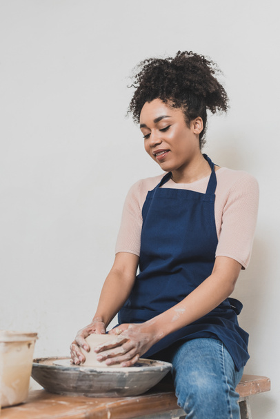 alegre joven afroamericana mujer sentada en el banco y modelando arcilla mojada en la rueda con las manos en cerámica - Foto, Imagen