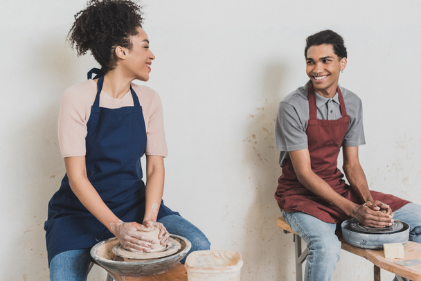 partial view of young african american couple looking at each other and modeling wet clay on wheels with hands in pottery - Photo, Image
