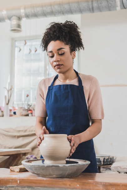 serious young african american woman modeling wet clay pot on wheel in pottery - Photo, Image