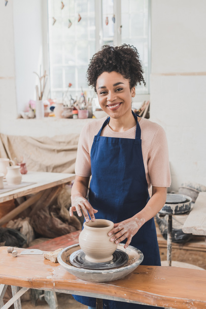 smiling young african american woman modeling wet clay pot on wheel in pottery - Foto, immagini