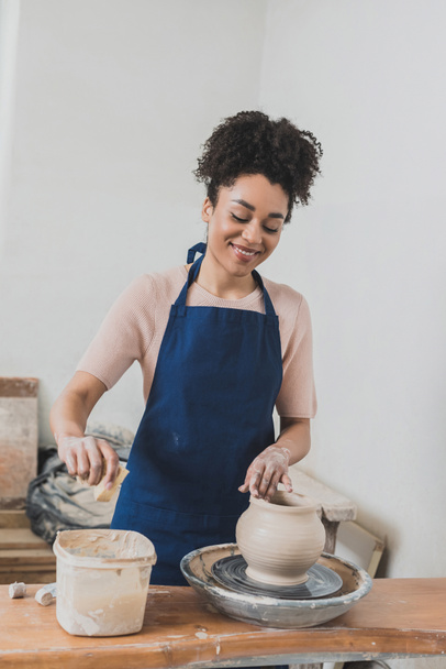 sonriente joven afroamericana mujer en delantal modelado mojado olla de arcilla en la rueda y apretando la esponja con la mano en cerámica - Foto, Imagen