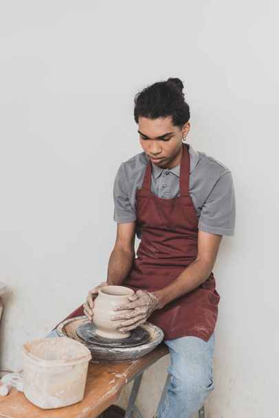 серьезный молодой африканский американец, моделирующий мокрый глиняный горшок на колесах с руками в керамике - Фото, изображение