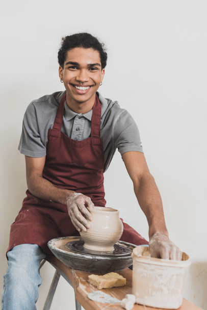 smiling young african american man modeling wet clay pot on wheel with hands in pottery - Photo, Image
