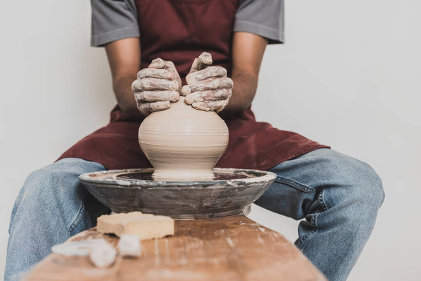 partial view of young african american man sitting on bench and shaping wet clay pot on wheel with hands in pottery - Fotoğraf, Görsel