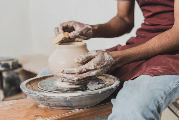 partial view of young african american man in jeans sitting on bench and shaping wet clay pot on wheel with sponge in pottery - Photo, Image