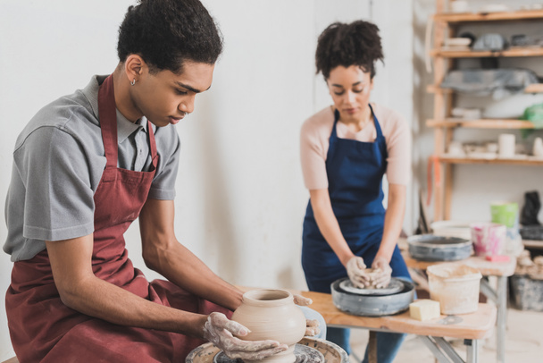 pareja afroamericana joven concentrada en ropa casual y delantales dando forma a ollas de arcilla húmeda en cerámica - Foto, imagen
