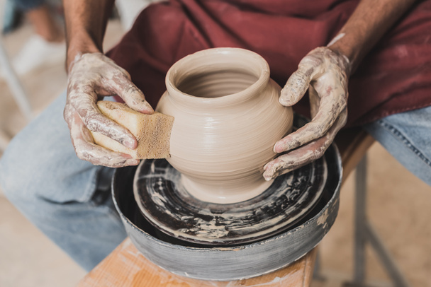 partial view of young african american man holding sponge and making wet clay pot on wheel in pottery - Photo, Image