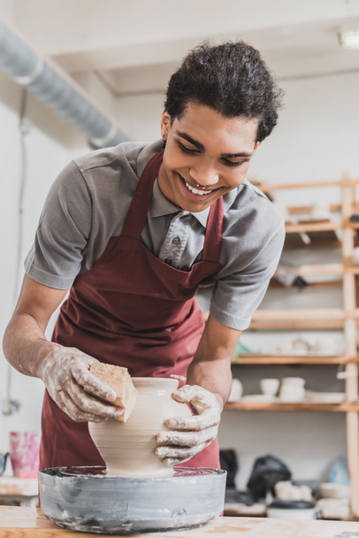 smiling young african american man making wet clay pot on wheel with sponge in pottery - Photo, Image