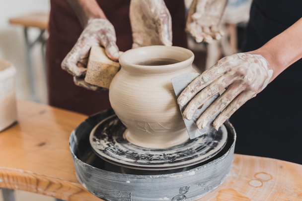 partial view of young african american couple shaping wet clay pot on wheel with hands and scraper in pottery - Photo, Image