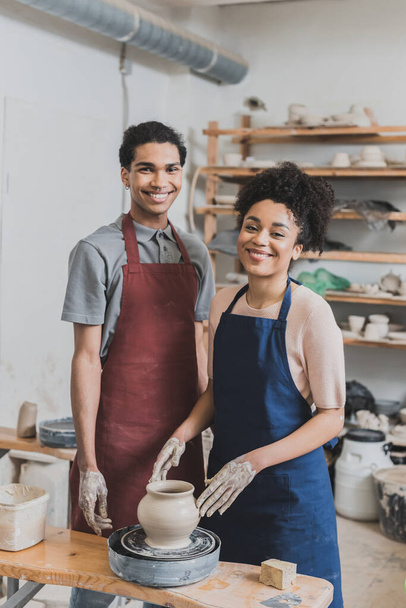 smiling young african american couple shaping wet clay pot on wheel in pottery - Photo, Image