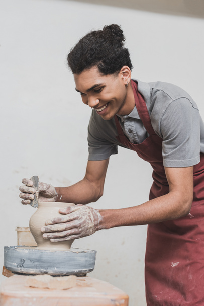 sonriente joven afroamericano hombre en delantal formando olla de arcilla húmeda con rascador en la rueda en cerámica - Foto, Imagen