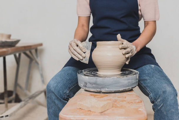 partial view of young african american woman sitting on bench and shaping wet clay pot with scraper in pottery - Fotografie, Obrázek