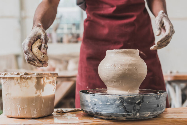 частичный вид молодого африканского американца в фартуке, который делает глиняный горшок на колесе и сжимает мокрую губку в керамике - Фото, изображение