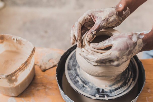 vista de cerca de manos afroamericanas masculinas esculpir olla de arcilla en la rueda en cerámica - Foto, imagen