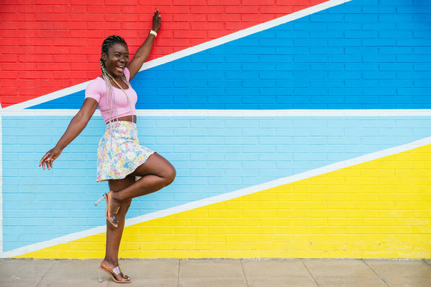 Веселая афро-американка в красочной юбке у разноцветной стены. Женщина с косичками, стоящая на одной ноге и вытянутыми руками, глядя в сторону. Концепция людей. - Фото, изображение