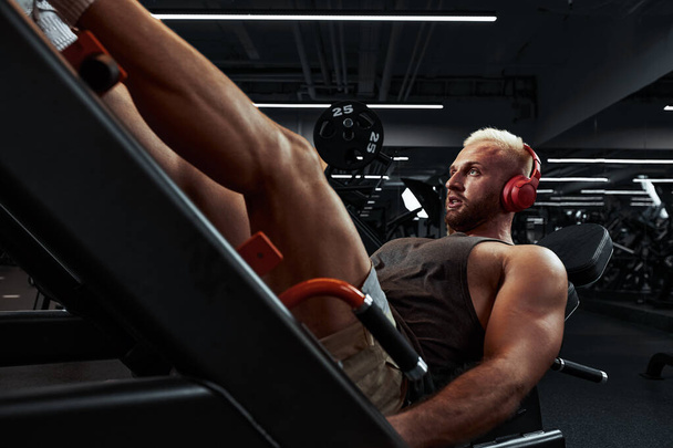 Человек тренирует ноги в тренажере. молодой человек делает упражнения на ногах в спортзале под музыку в наушниках - Фото, изображение