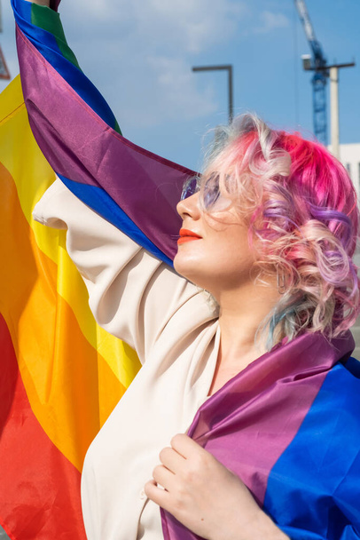 Caucasian woman with curly colored hair holding lgbt flag. Lesbian woman holding a rainbow flag outdoors - Фото, изображение