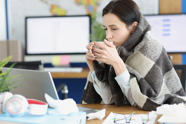 Sick young woman sits in warm blanket with cup at workplace - Zdjęcie, obraz