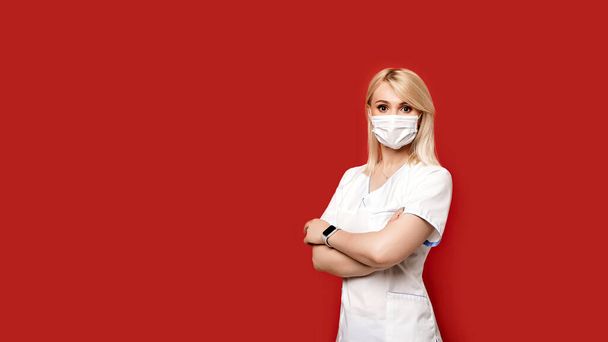 Confident female doctor wearing protective face mask standing on red background. Young woman intern in medical clothes posing. - Fotografie, Obrázek