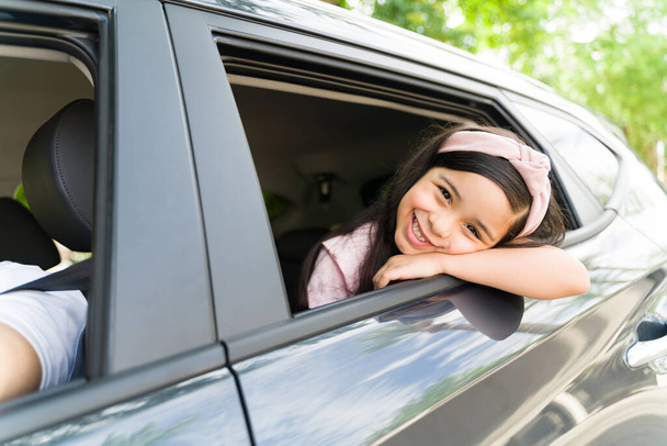 Adorable fille élémentaire souriant et se sentant mignon tout en regardant la caméra. Petite fille attendant sa famille dans la voiture pour partir en voyage de vacances - Photo, image