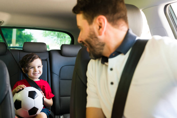 Homme attentionné et père regardant en arrière son petit garçon dans la voiture et parlant tout en le conduisant à l'entraînement de football et en faisant des courses - Photo, image