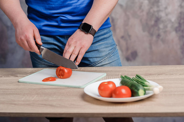 A man cuts a red tomato on a board. Cucumbers and tomatoes in a white plate - Photo, Image