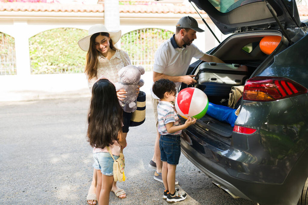 Happy beautiful family putting suitcases, floaters and a beach ball on the car. Latin parents talking with their son and daughter about summer vacations - Fotó, kép