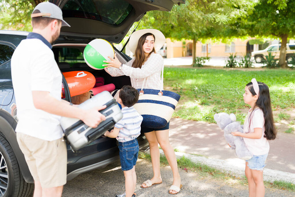 Привлекательная молодая женщина разговаривает со своими маленькими детьми об их летних каникулах и семейной поездке, упаковывая свои вещи в машину  - Фото, изображение
