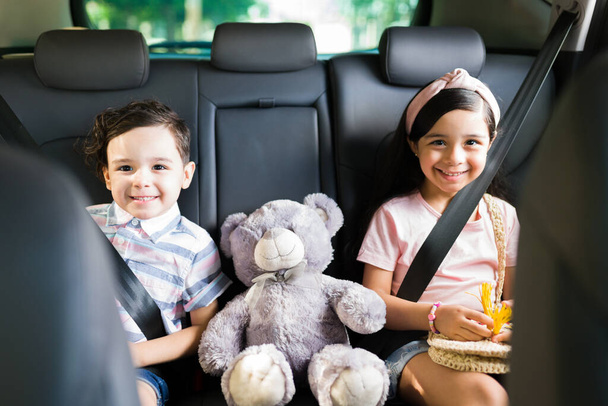 ¡Emocionados por nuestras vacaciones familiares! Retrato de dos lindos hermanos felices sentados con un oso de peluche en el coche y sonriendo durante un viaje por carretera - Foto, Imagen