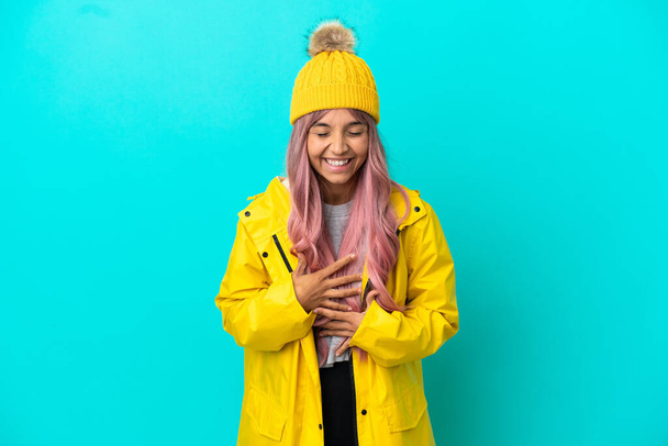 Young woman with pink hair wearing a rainproof coat isolated on blue background smiling a lot - Photo, Image