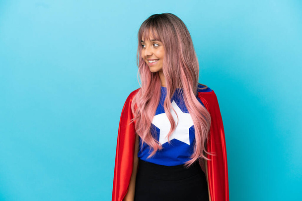 Mladý superhrdina žena s růžovými vlasy izolované na modrém pozadí při pohledu do strany a usmívá - Fotografie, Obrázek