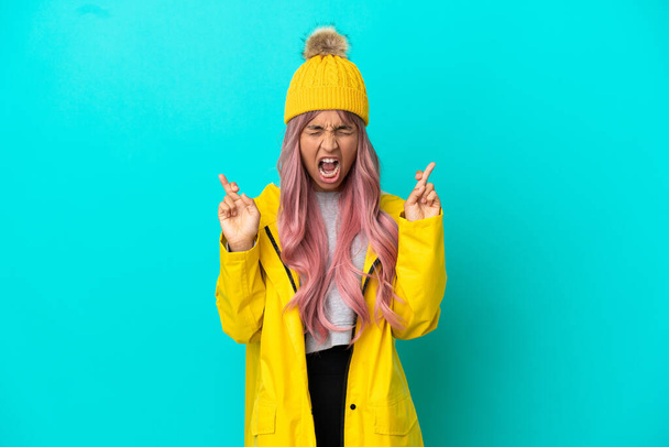 Young woman with pink hair wearing a rainproof coat isolated on blue background with fingers crossing - Foto, immagini