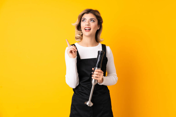 Girl using hand blender isolated on yellow background intending to realizes the solution while lifting a finger up - Photo, Image