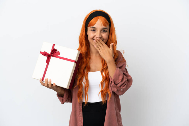 Turuncu saçlı genç bir kadın beyaz arka planda izole edilmiş bir hediye tutuyor mutlu ve ağzı kapalı gülümsüyor. - Fotoğraf, Görsel