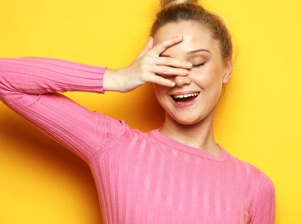An emotional young woman in a pink jumper smiles and touches her face. Happiness and joy. Portrait on a yellow background. - Fotografie, Obrázek