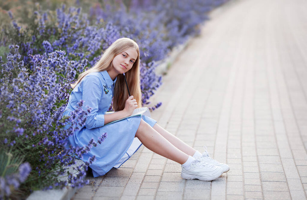Chica adolescente con pluma escribe en un cuaderno, sentado en el parque en flores de lavanda azul fuera. concepto de educación y aprendizaje - estudiante feliz haciendo la tarea - Foto, Imagen