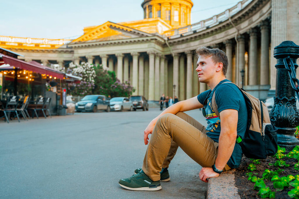 A young man sits on the sidewalk in front of the Kazan Cathedral in summer. Saint Petersburg, Russia - 05 Apr 2021 - Photo, Image