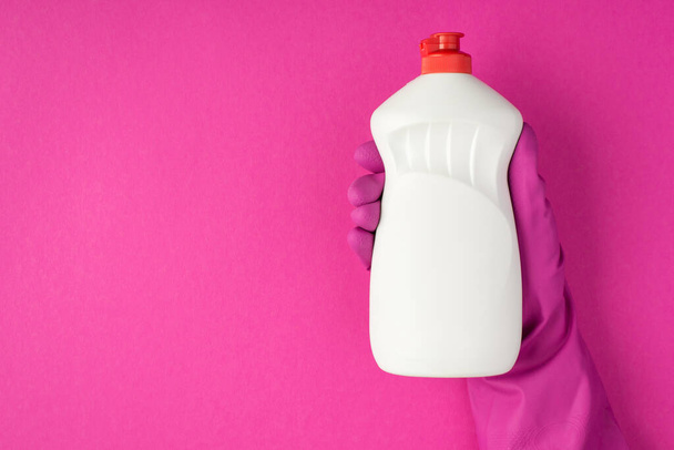 Φωτογραφία του χεριού σε ροζ γάντι που κρατά λευκό τζελ απορρυπαντικό μπουκάλι χωρίς ετικέτα σε απομονωμένο ροζ φόντο με κενό χώρο - Φωτογραφία, εικόνα