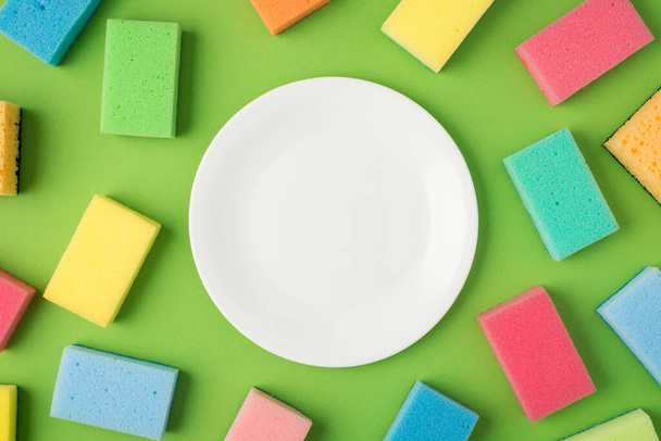 Top view photo of white clean plate in the middle and multicolor scouring pads on isolated green background with copyspace - Photo, Image