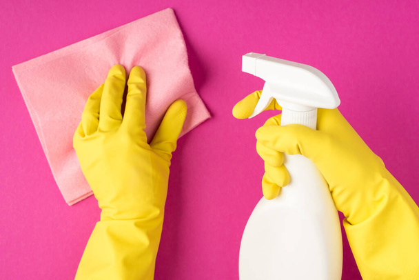 Vista superior de la foto de las manos en guantes amarillos con trapo de viscosa rosa pastel y el uso de botella de detergente en aerosol sin etiqueta sobre fondo rosa aislado con copyspace - Foto, Imagen